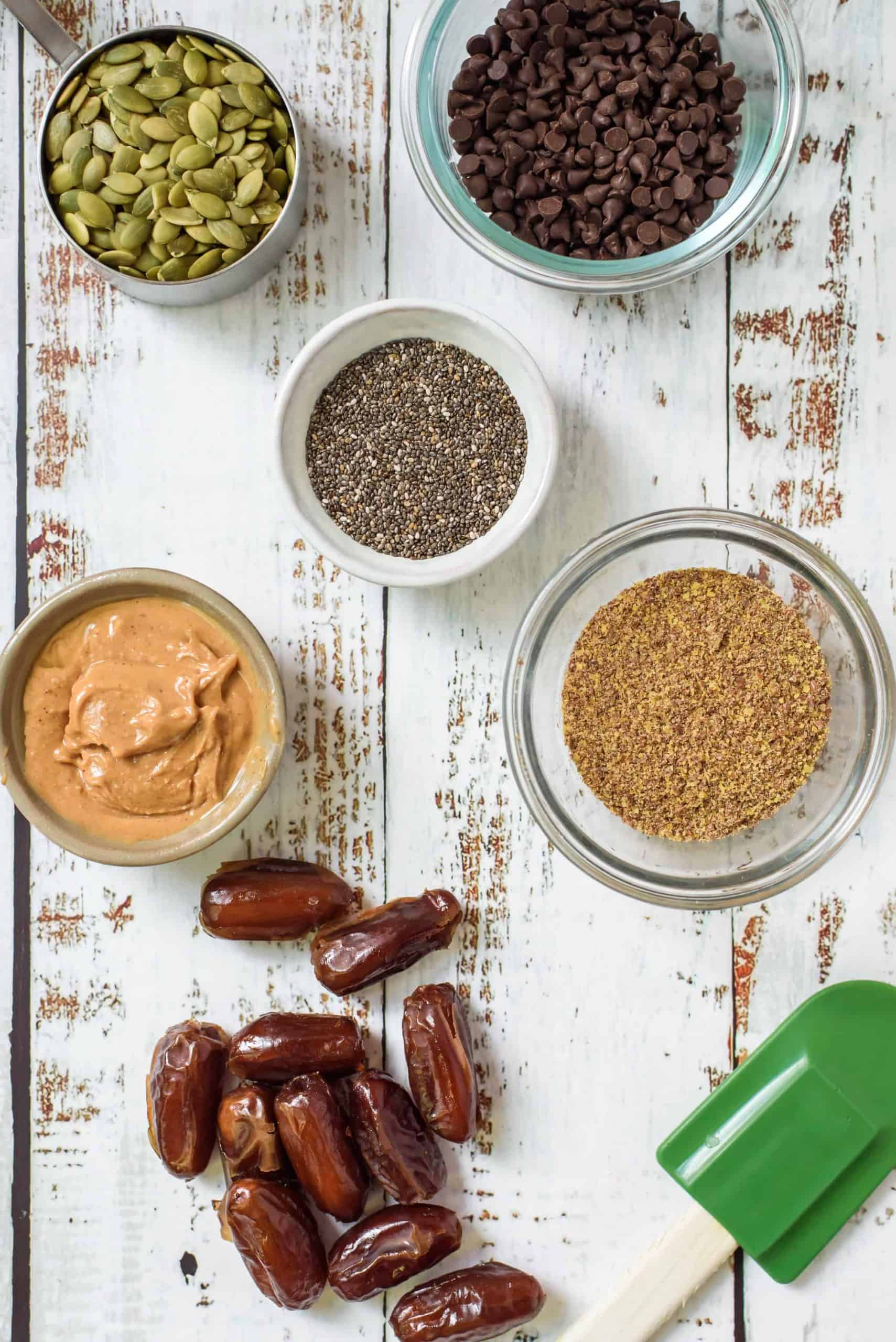 Overhead shot of dates, peanut butter, flax seed, chia seed, pumpkin seed and mini chocolate chips on a wood board with a green spatula.
