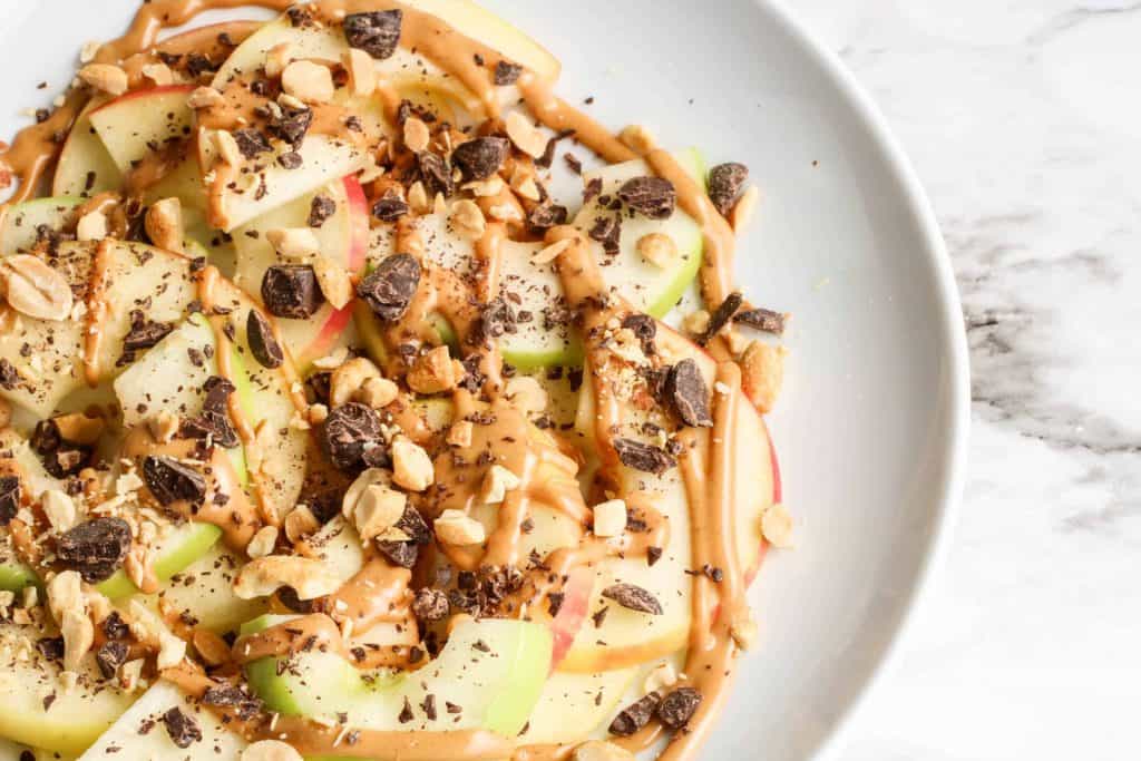 Overhead view of apples with peanut butter, nuts and chocolate chips on them on a white plate.