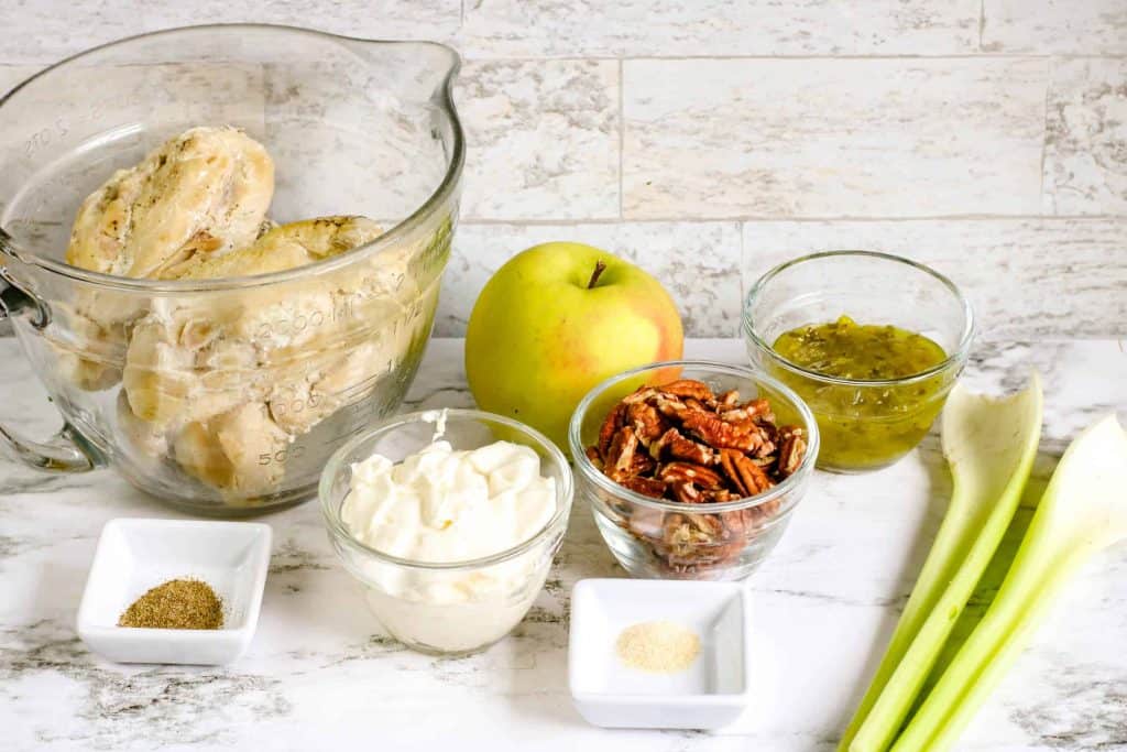 Cooked chicken breast, mayonnaise, pecans, apple, relish, and celery on a marble background.