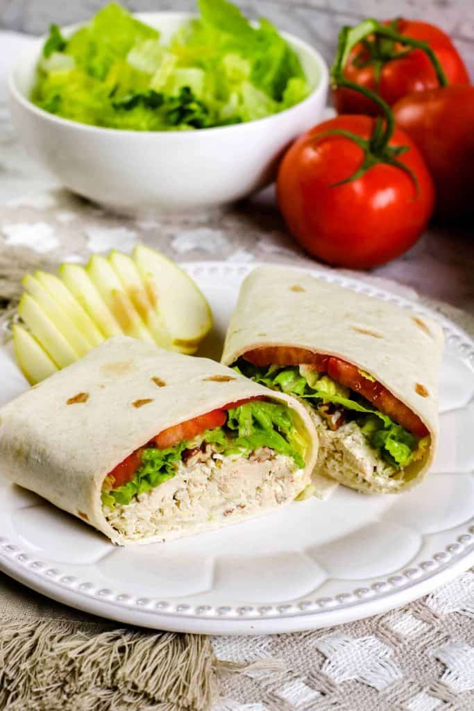 Chicken salad wrap on a white plate with sliced apples on the side and a bowl of lettuce and two red tomatoes in the background.