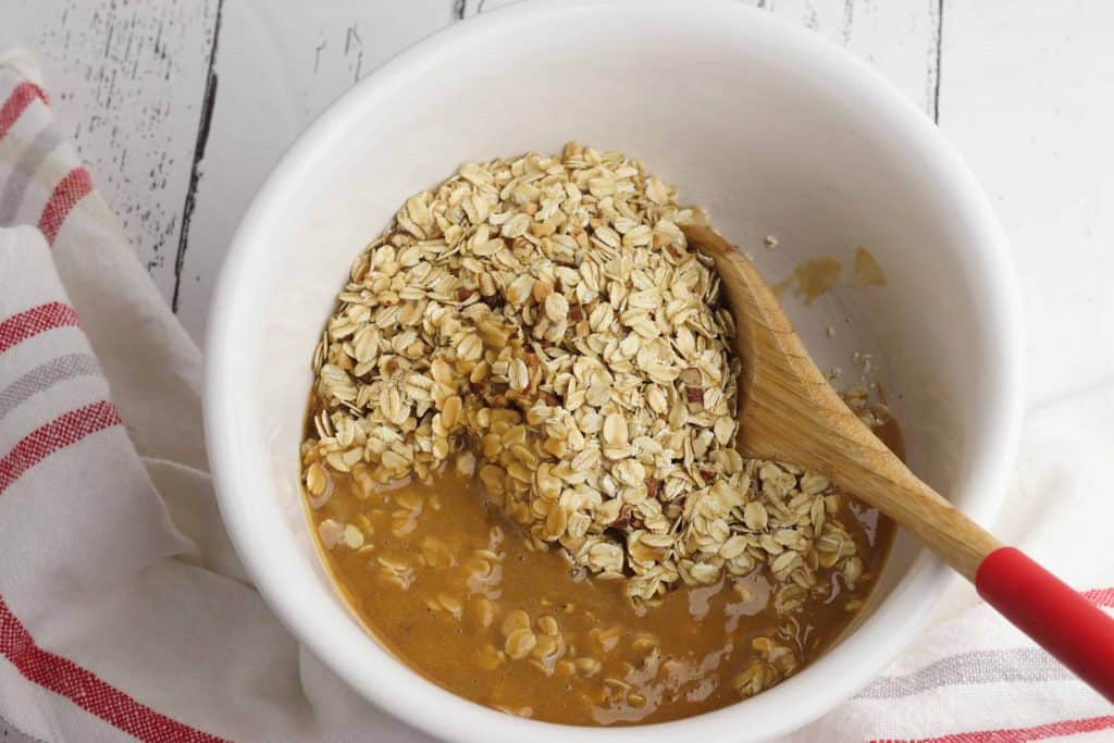 Rolled oats with melted peanut butter mixture on top of them in a white ceramic bowl with a wood spoon in the bowl.