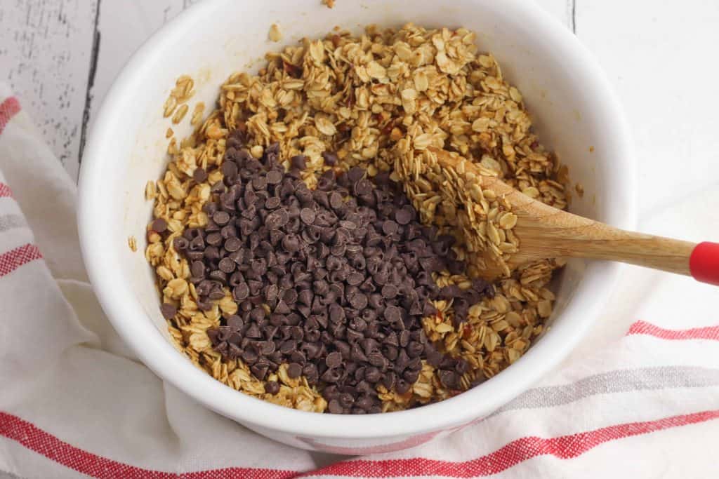 Rolled oats and chocolate chips in a white ceramic bowl with a wooden spoon in the center.