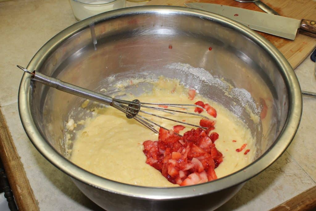 Bread dough a stainless steel bowl with whisk and cut up strawberries on top.