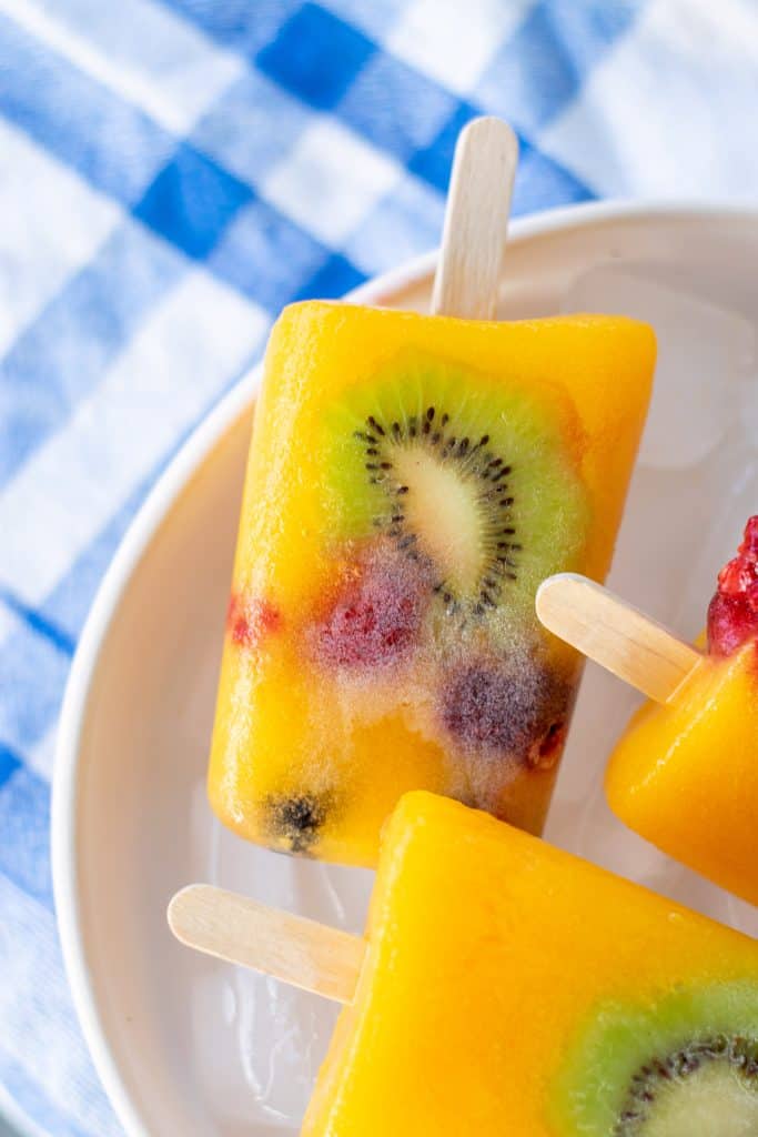Closeup of Kiwi mango popsicles in a bowl of ice on blue checked table cloth.