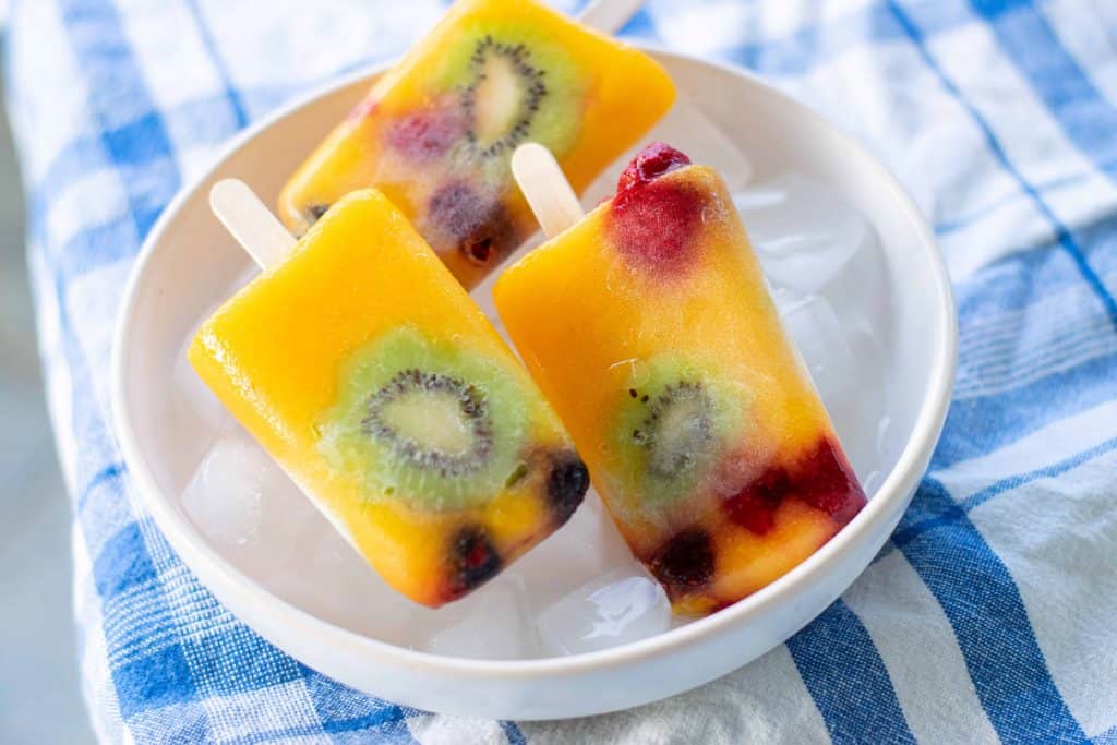 Kiwi Mango popsicles in white bowl with ice on blue checked tablecloth.