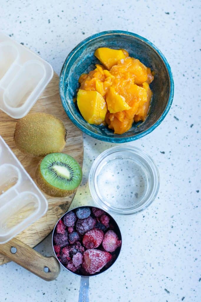 Mango chunks, frozen berries, kiwi, popsicles molds and glass bowl of water.