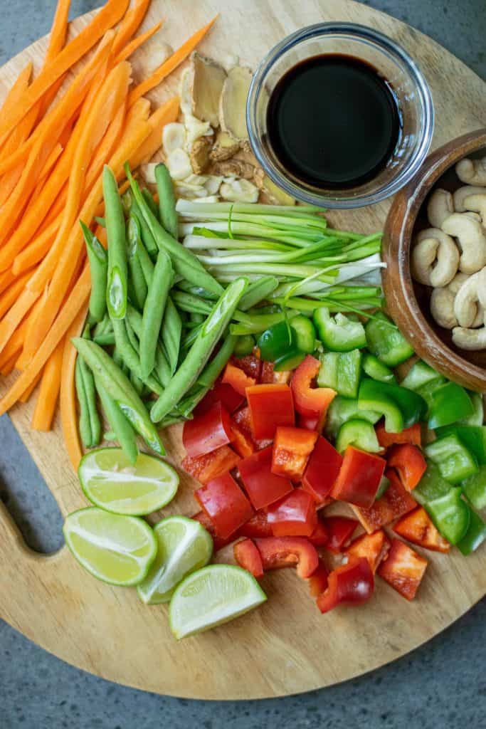 Sliced carrots, green beans, bell peppers, onions, lime slices on cutting board with a bowl of Kung Pao sauce and cashews on the side.