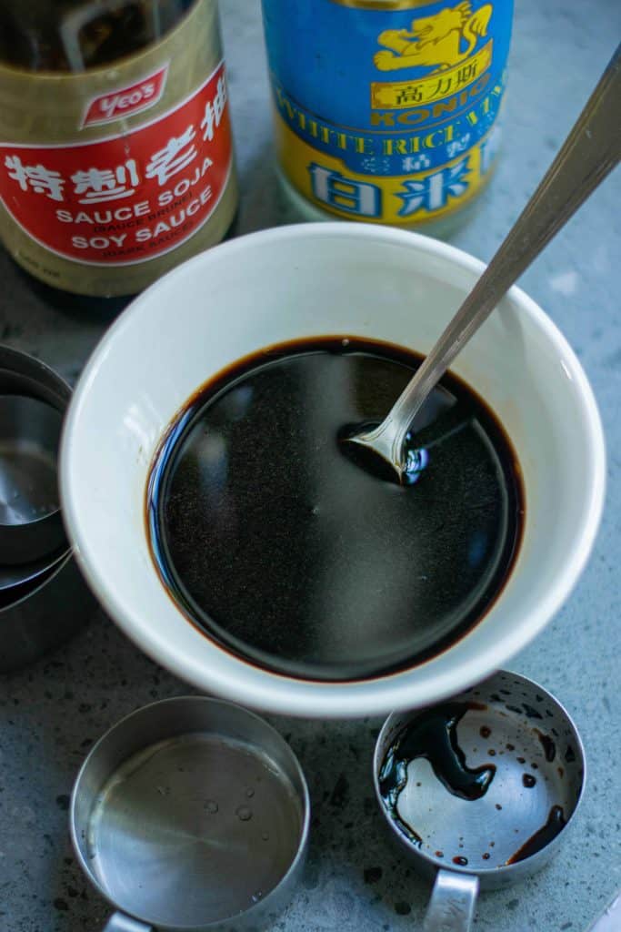 Soy sauce in a white bowl with spoon in it and measuring cups and bottles around it.