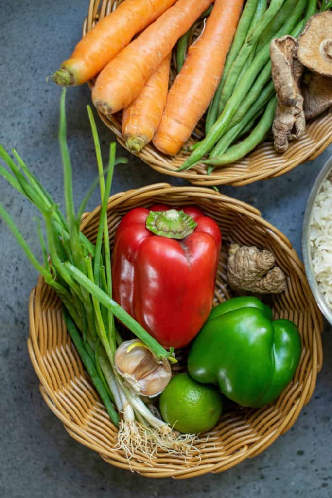 Bell peppers, onions, garlic and ginger in a wicker basket and carrots and green beans and mushrooms in another wicker basket. White rice peaking in the side.