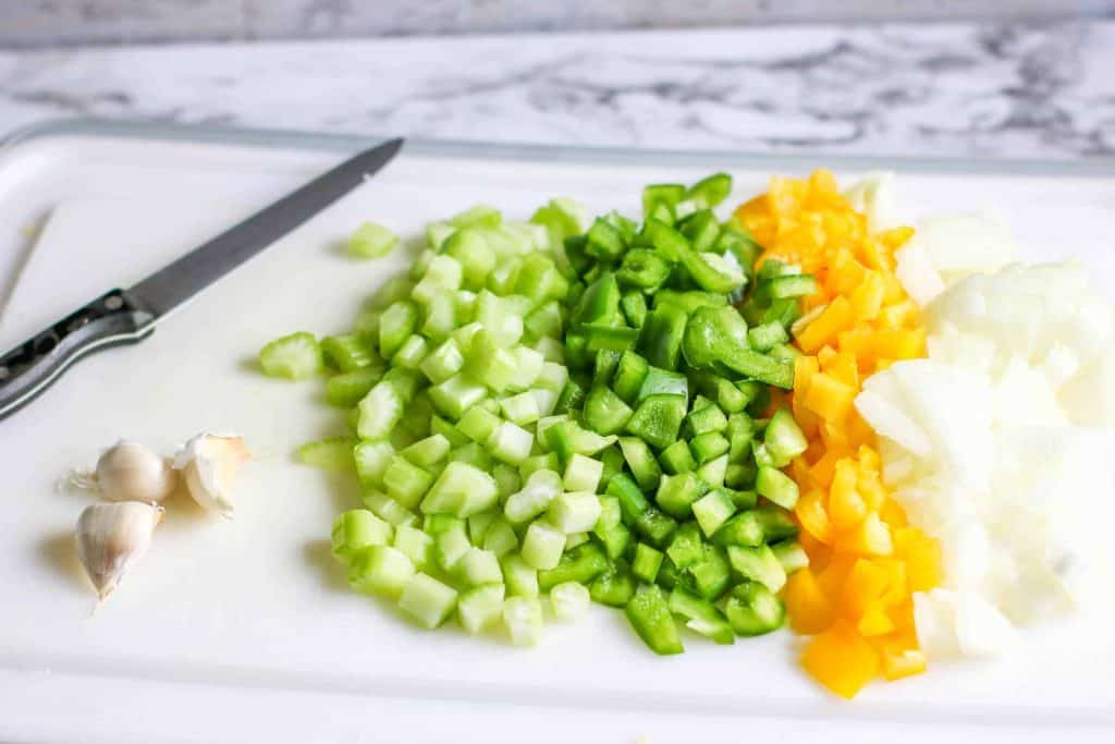 Diced celery, green and yellow bell pepper, onions and garlic on a cutting board with a knife on the side.