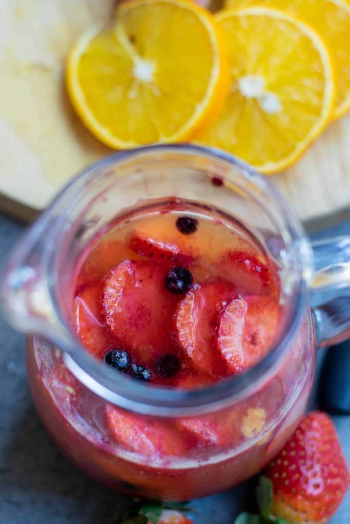 Overhead photo of Sangria with strawberry slices and blueberries, orange slices on a cutting board on the side.