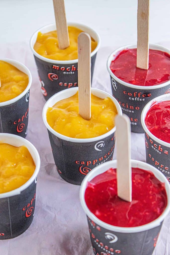 Puréed mango and raspberry frozen yogurt cups with popsicle sticks in the center.