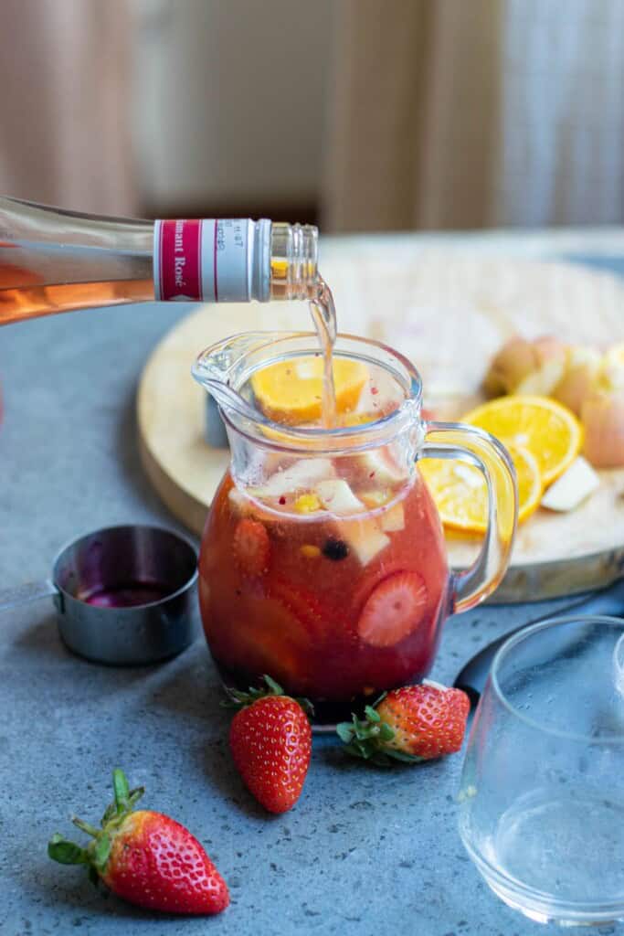 Pouring Ros&eacute; wine from a bottle into a glass pitcher of sangria with vodka with fresh strawberries oranges and blueberries