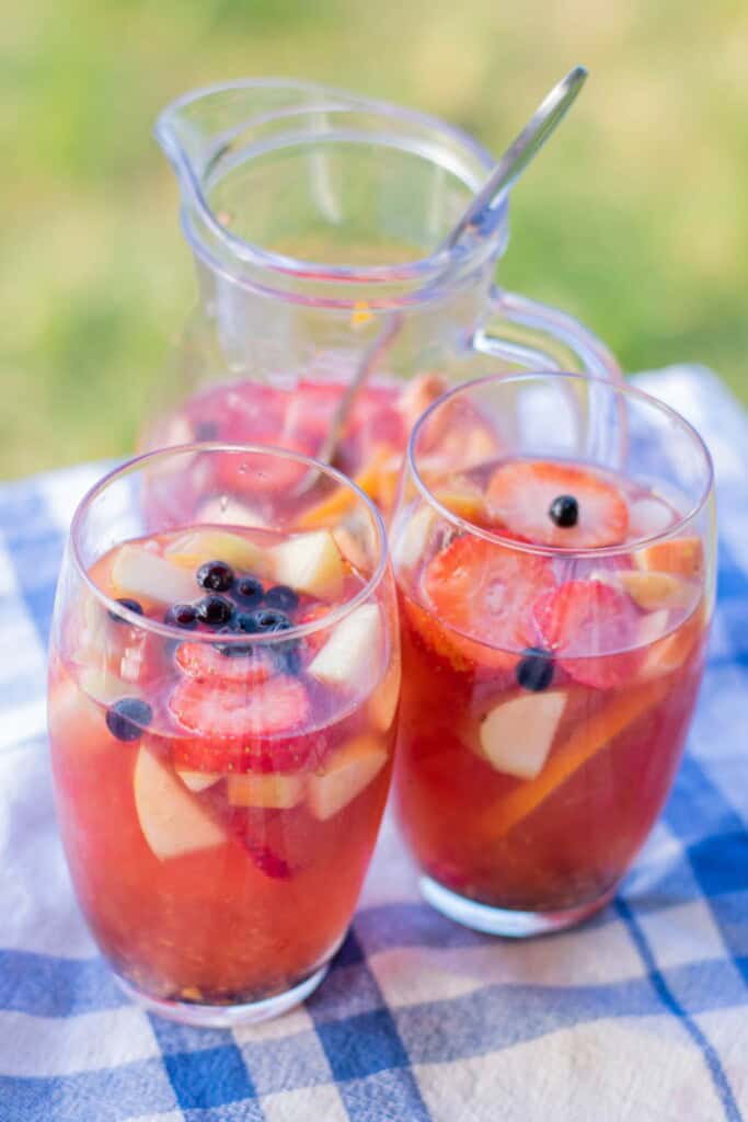 Two tall glasses and a glass pitcher of Ros&eacute; sangria with vodka garnished with fresh strawberries blueberries and apple chunks on a blue checked tablecloth outdoors