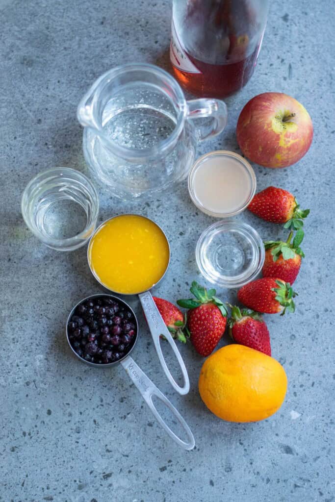 Ingredients for sangria with vodka including Ros&eacute; wine vodka mango juice sparkling water fresh strawberries apple orange and frozen blueberries on a gray countertop