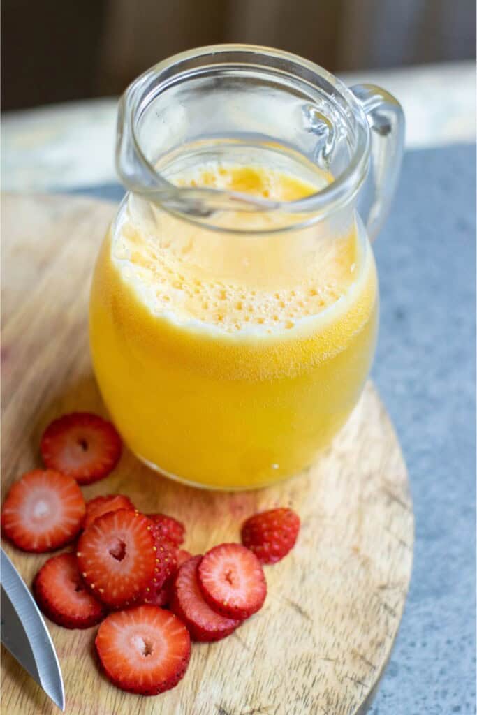 Sliced fresh strawberries on a wooden cutting board next to a glass pitcher of mango juice and sparkling water for sangria with vodka