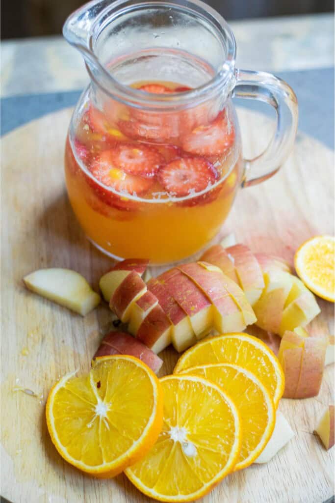 Glass pitcher with mango juice and sliced strawberries surrounded by diced apple and orange slices on a wooden cutting board for vodka sangria