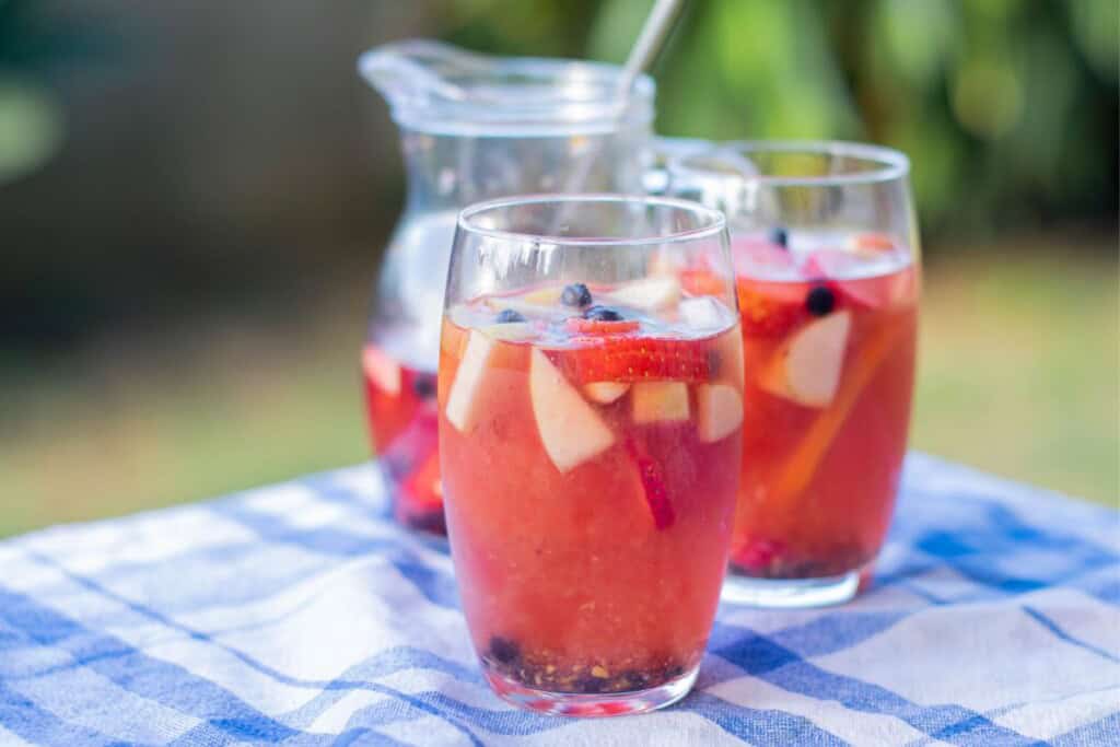 Two glasses of vodka sangria with Ros&eacute; wine garnished with strawberries blueberries and apple chunks beside a serving pitcher on a blue checked tablecloth outdoors