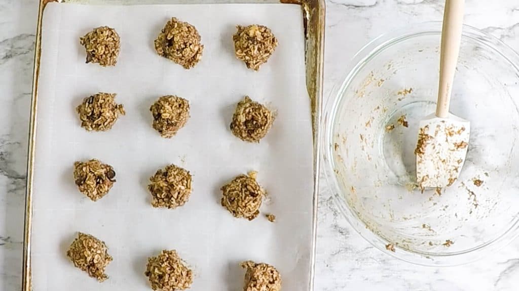 Cookie dough balls on a baking pan lined with parchment bowl. Empty glass bowl with spatula in it on right hand side.