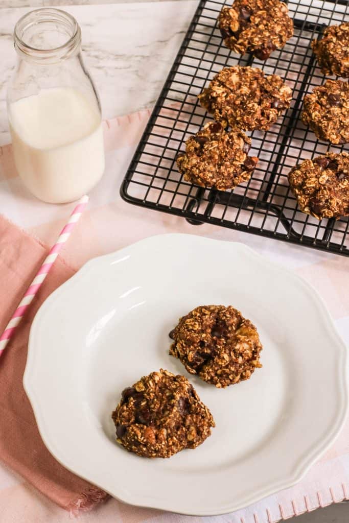 Banana Bread Cookies on a wire cooling rack in upper right hand corner and 2 cookies on a white plate. A glass of milk in the upper left hand corner.