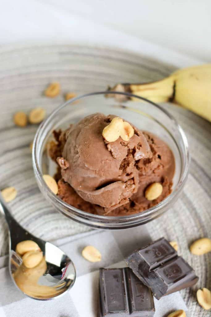 Chocolate Nice Cream in a glass bowl with peanuts on top and a banana peaking in on the side and a silver spoon and chocolate bar pieces.