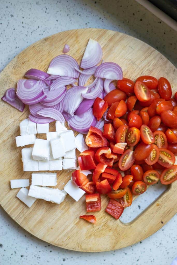 Sliced red onion, cubed feta cheese and halved cherry tomatoes on a wood cutting board.