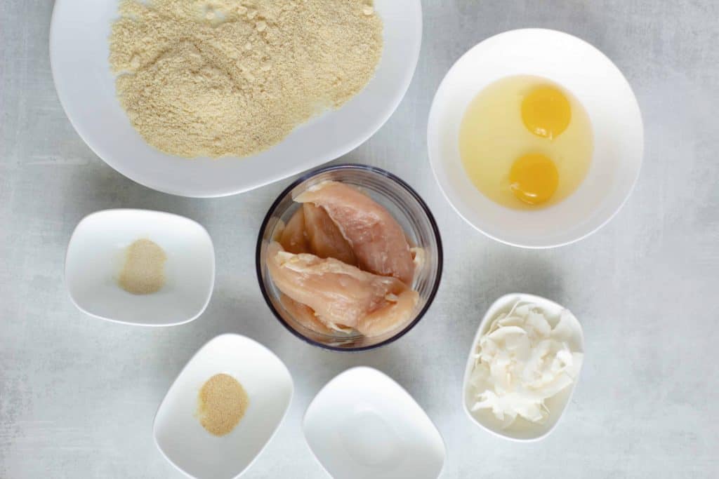 Chicken Tenders, eggs, coconut chips and breading in white bowls on a white background.