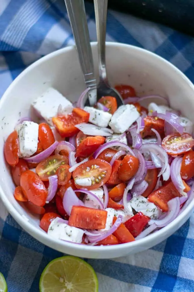 Mediterranean Tomato Salad in a white serving bowl with 2 serving spoons on a blue checked table cloth.