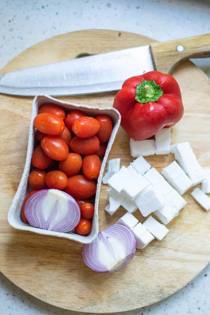 Cherry tomatoes and ½ red onion in a carton on a cutting board along with red bell pepper, cubed feta cheese and half red onion on a round wood cutting board with a chef's knife at the top.
