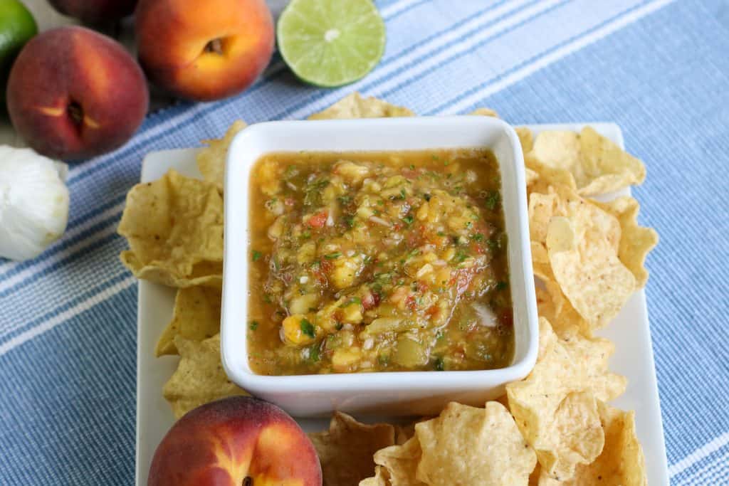 Fresh peach salsa in a square white bowl dish with tortilla chips around it on a blue table cloth with fresh peaches in the corner and on the side and half of a lime.