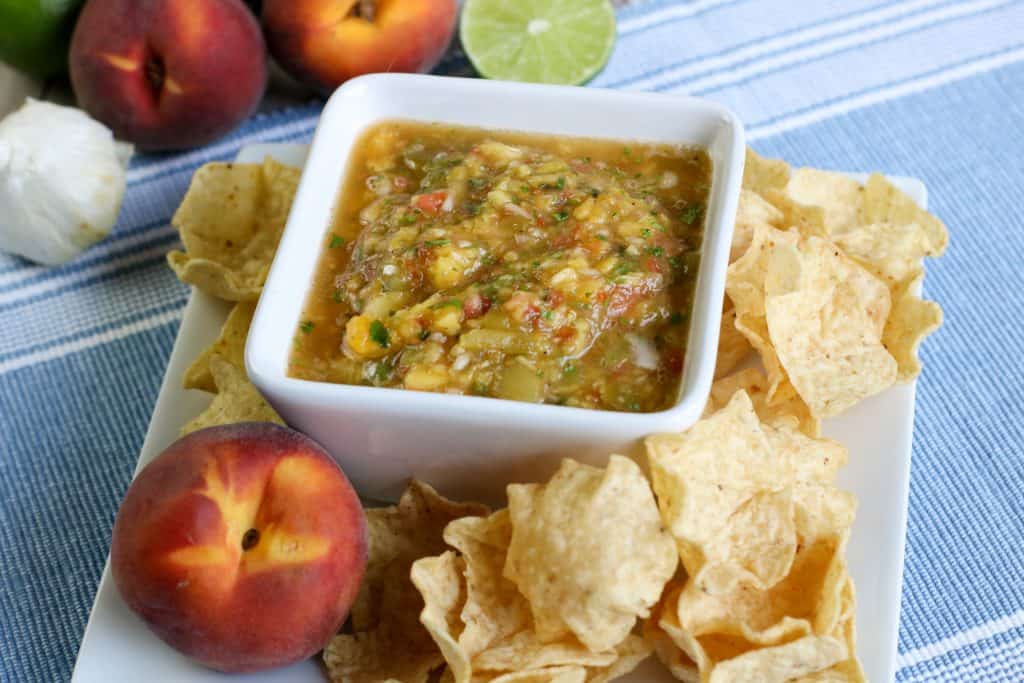 peach salsa in a white square bowl on a white square platter with chips and whole peaches around it.