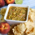 peach salsa in a white square bowl on a white square platter with chips and whole peaches around it.