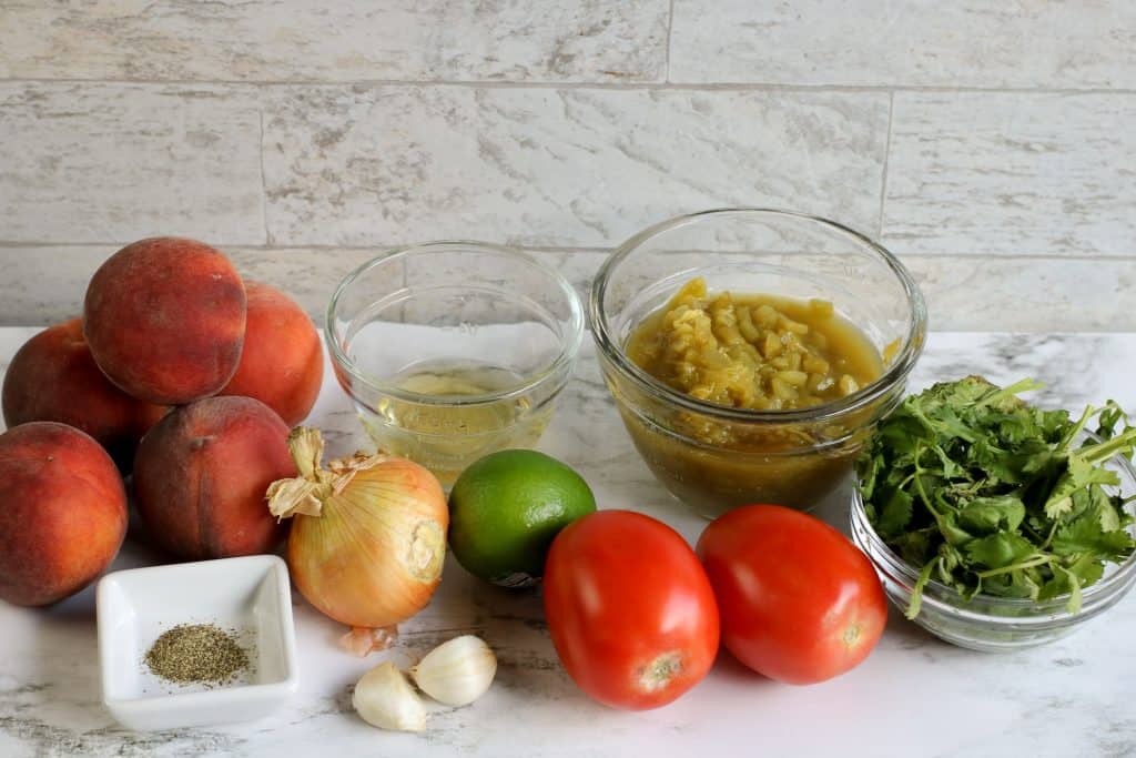 Fresh peaches, apple cider vinegar, tomatoes, cilantro, lime garlic, onion, salt and pepper and diced green chiles on a white marble background.
