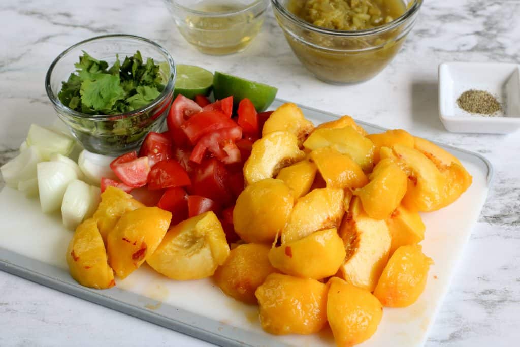 Diced fresh peaches, diced tomatoes, diced onions, cilantro and lime sliced in half on a cutting board.