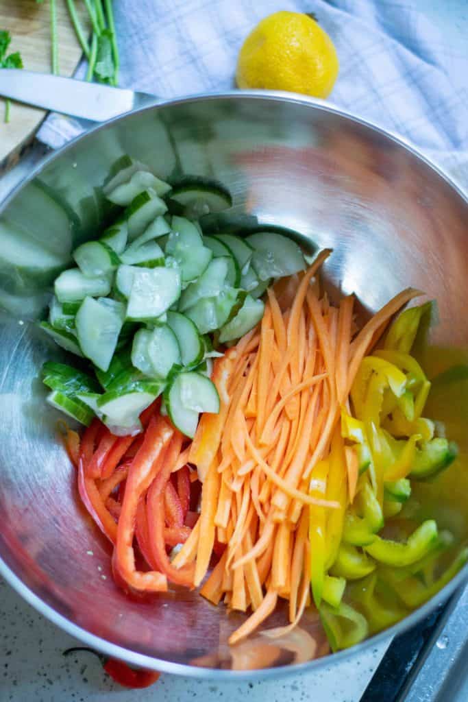 Overhead shot of cucumber, bell pepper strips, carrot strips and yellow bell pepper strips in a stainless steel bowl.