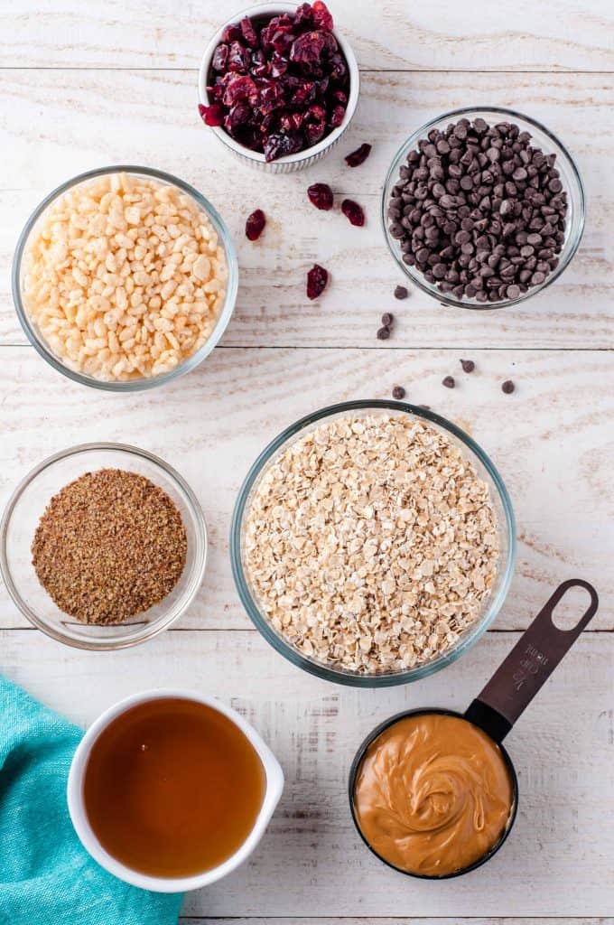 Oats, peanut butter, agave syrup, chia seeds, chocolate chips, and dried cranberries in small bowls on a white wood background with a blue napkin on bottom corner.