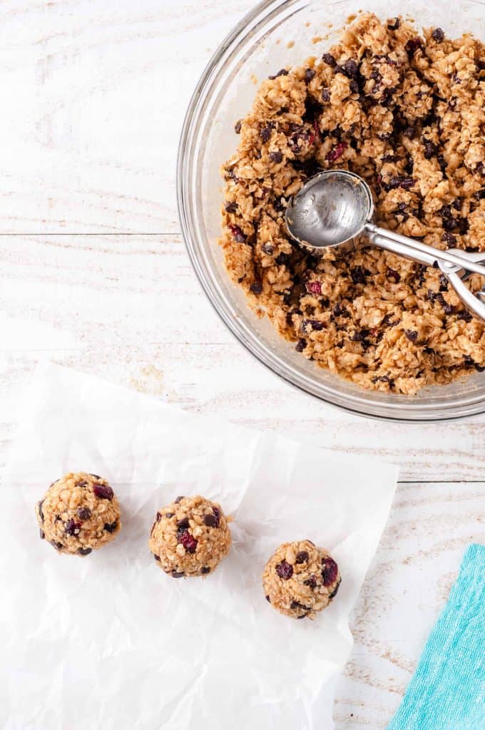 Vegan Protein Ball Mixture in glass bowl with cookie scoop in it. 3 vegan protein balls on parchment paper below it.