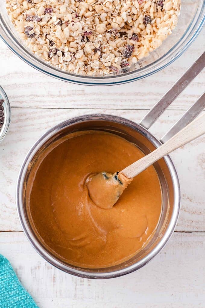 Melted peanut butter in a sauce pan with a wood spoon in it. Large bowl of oats with chocolate chips in it above saucepan.