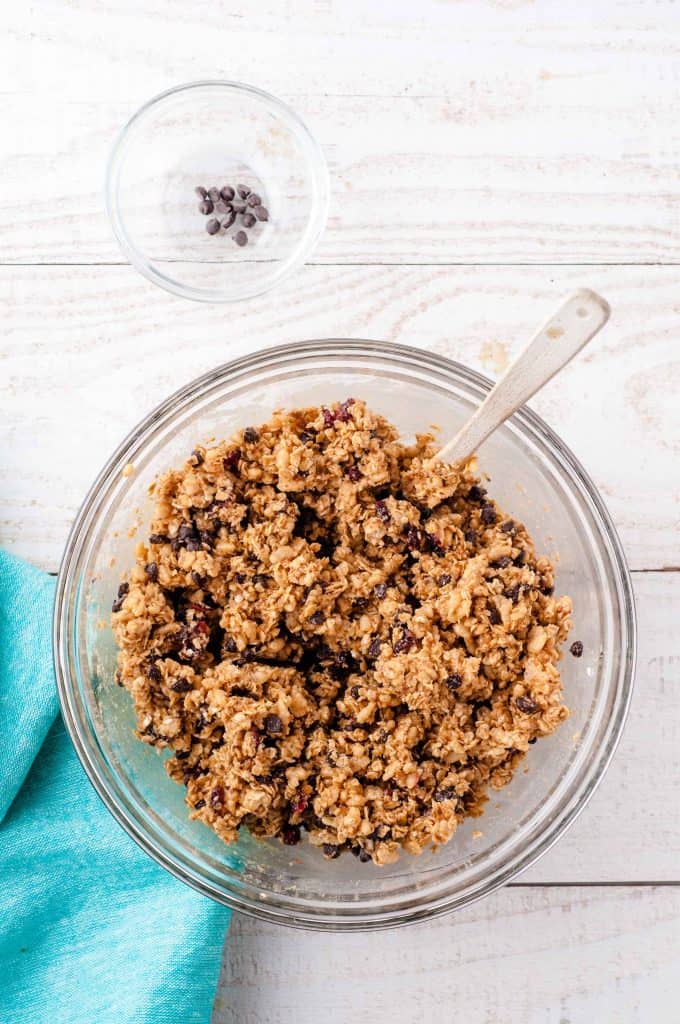 Vegan Protein Ball Mixture in a glass bowl with a blue napkin in bottom left corner and small glass bowl with a few chocolate chips in it above large glass bowl with mixture.