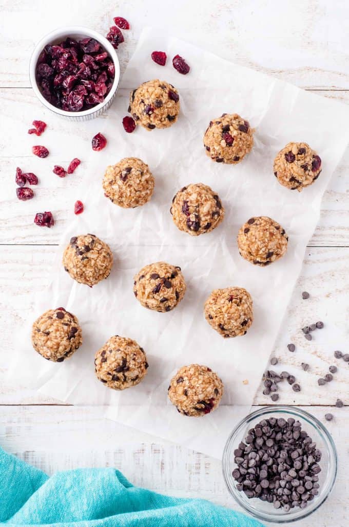 12 vegan protein balls on parchment paper with dried cranberries in a small white bowl above it and chocolate chips in a small glass bowl below it with a blue napkin peaking out of right bottom corner.