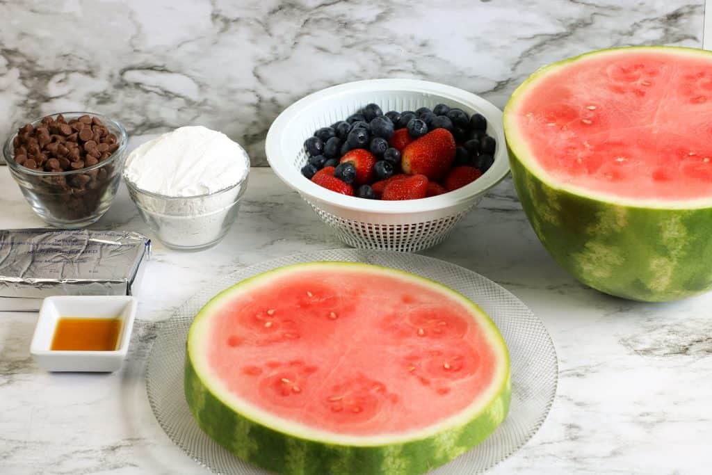 Large slice of watermelon on a glass plate, bowl of berries, bowl of powdered sugar, milk chocolate chips, cream cheese, and vanilla.