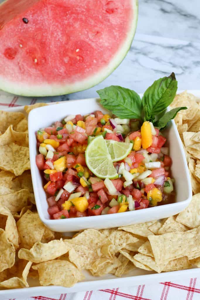 Watermelon salsa with lime slices on top and mint on the side, surrounded by tortilla chips with watermelon above it on a marble background.