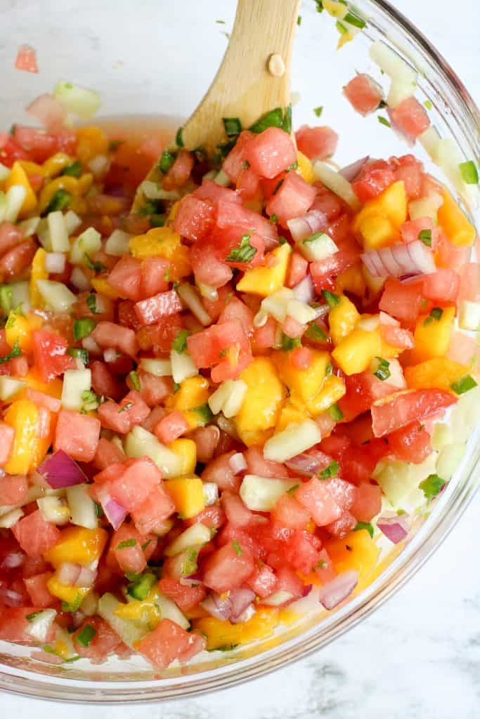 Fresh watermelon salsa being stirred with a wooden spoon in a large glass bowl.