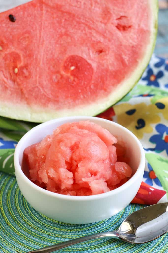Watermelon Sorbet in a white bowl with a spoon by the side and a slice of watermelon behind it.