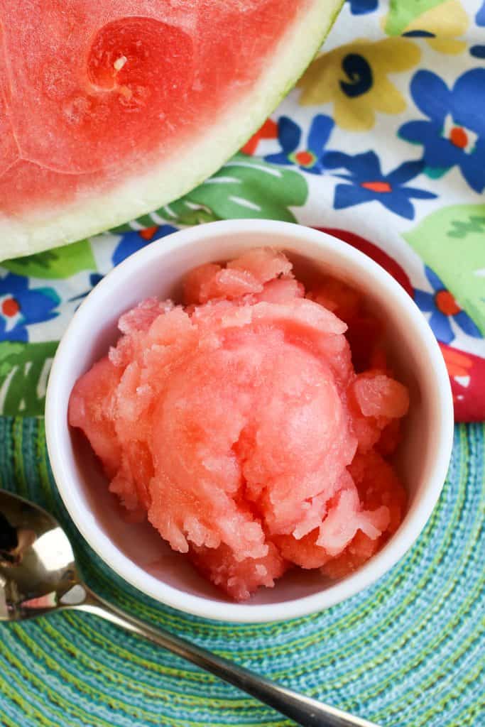 Overhead shot of Watermelon Sorbet on a teal background with a silver spoon on the side and a flowered napkin in the right corner and a slice of fresh watermelon on the there corner.