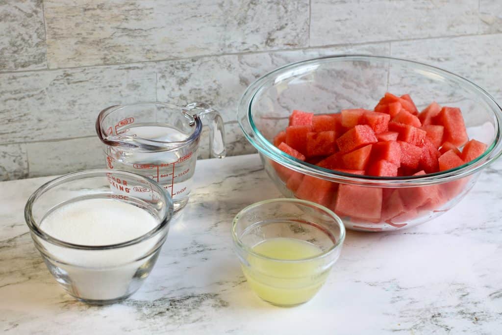 Water in a measuring cup, sugar in a glass bowl, lemon juice in a glass bowl and watermelon in a glass bowl on a marble background.