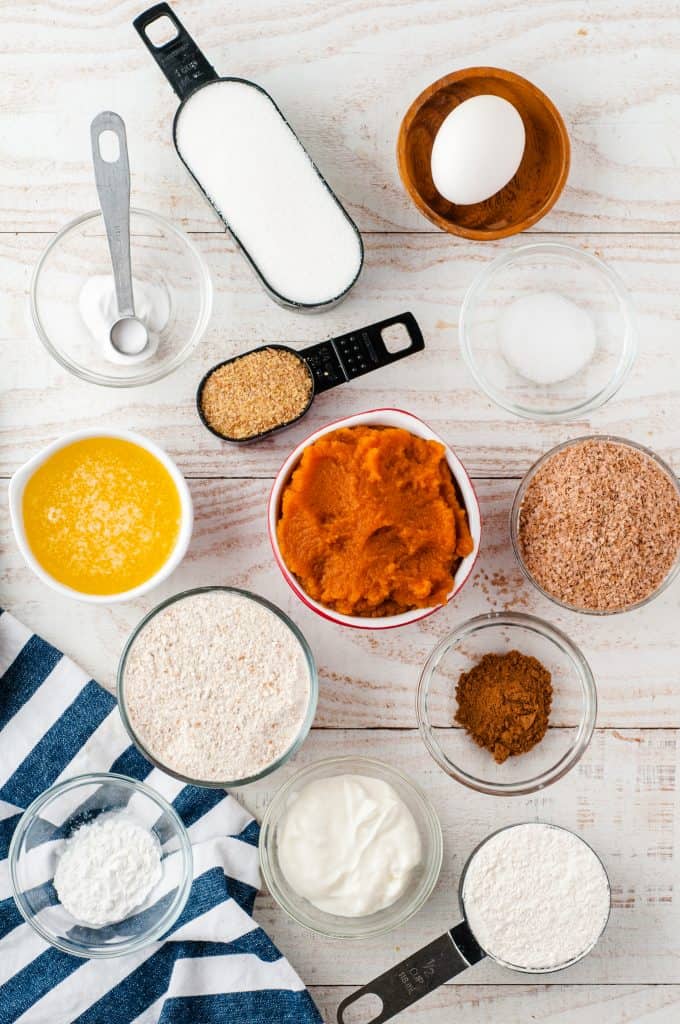 flour in a measuring cup, egg in a wood bowl, brown sugar in a measuring spoon, melted butter in a white bowl, brown sugar in a glass bowl, cinnamon in a glass bowl, wheat flour in a measuring cup, wheat germ in a glass bowl and pumpkin puree in a white bowl.