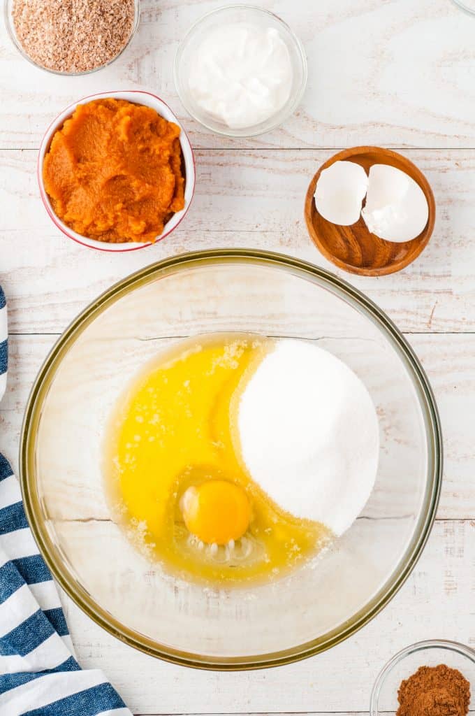 Eggs, melted butter and flour in a glass bowl with pumpkin puree in a bowl on the side and egg shell in a bowl in the side.