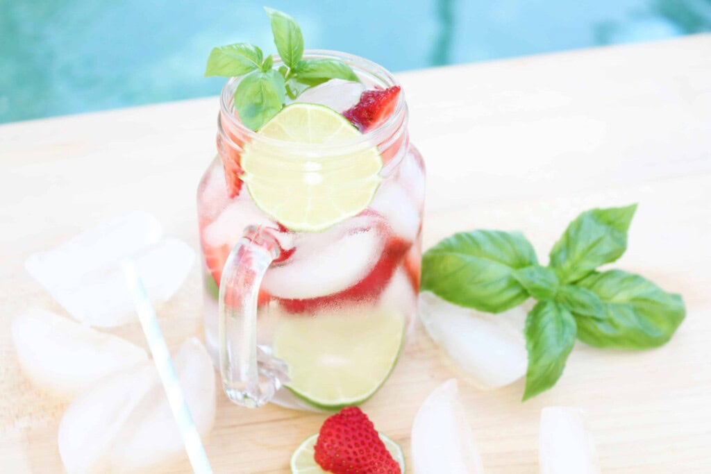 Strawberry Basil Cocktail with ice cubes, fresh basil and strawberry around glass setting beside a pool.