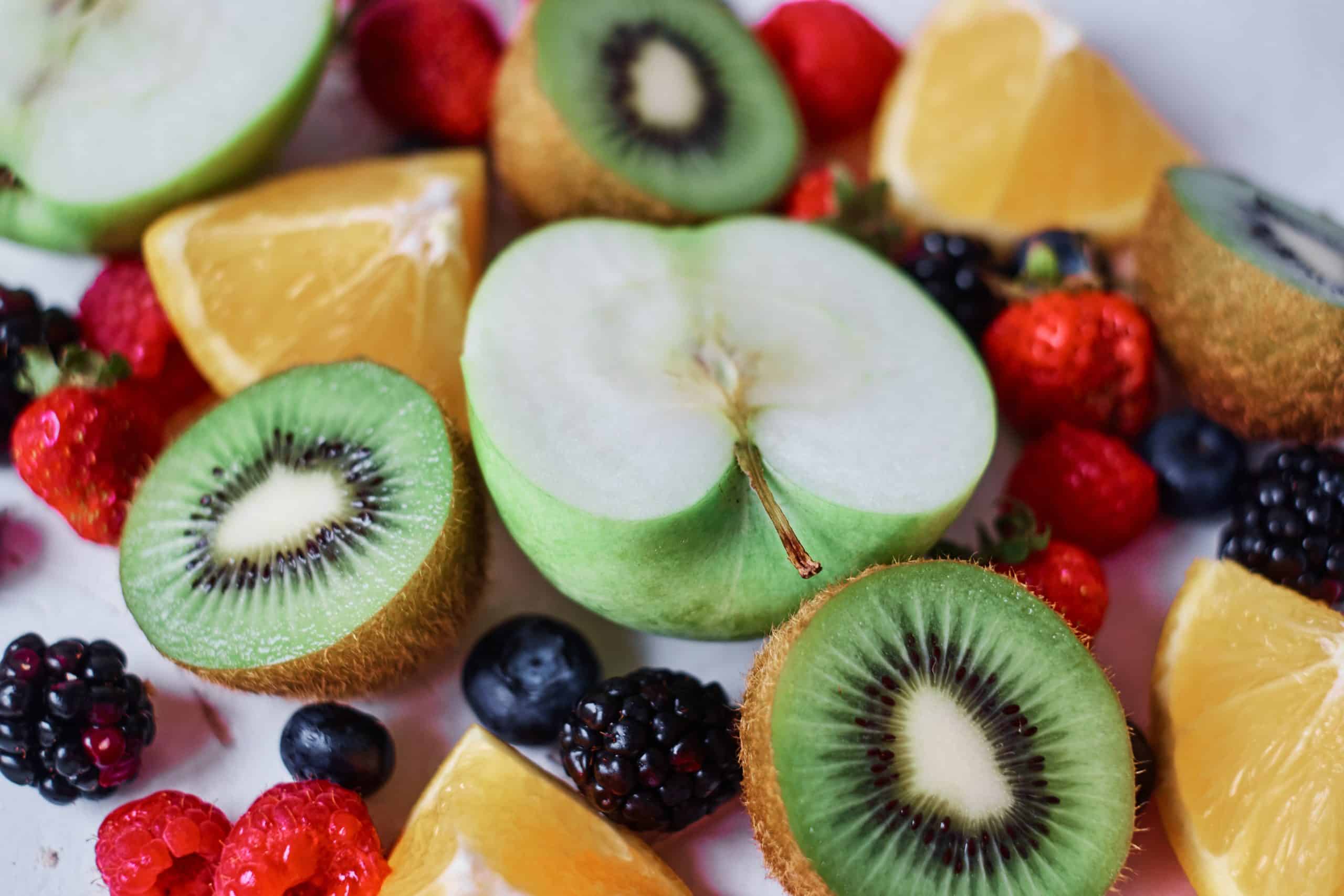 Kiwis and green apples sliced in half, with quartered oranges, blackberries, blueberries and raspberries on a white background.